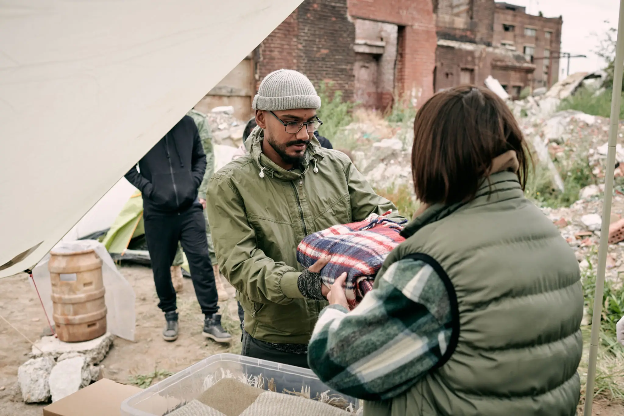 Rear view of social worker in warm vest giving plaid to middle-eastern migrant in glasses and hat outdoors