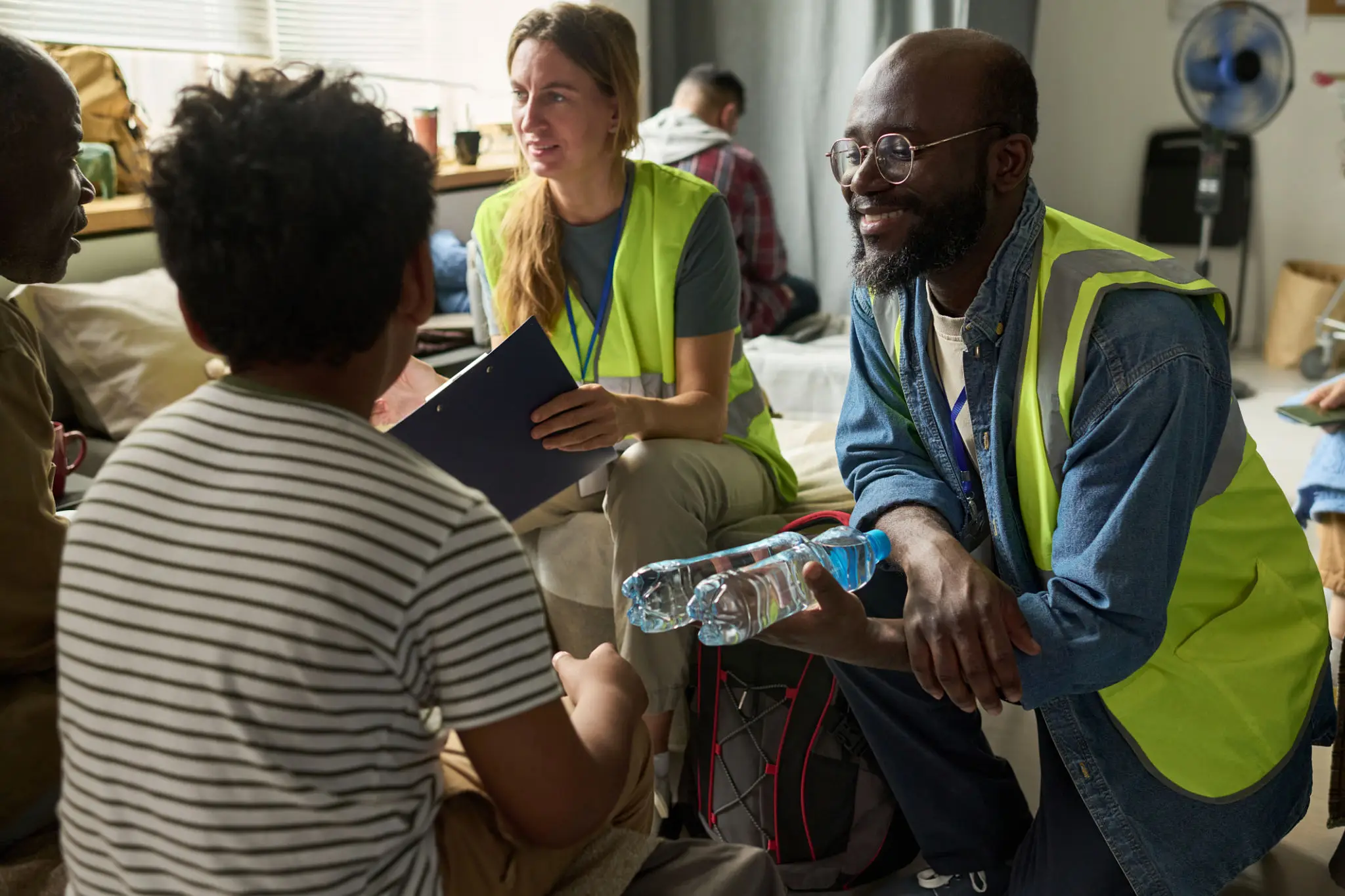 Medium shot of diverse aid workers distributing water and coordinating support with displaced adults in shelter, representing humanitarian relief. Ideal for nonprofit campaigns and crisis response messaging